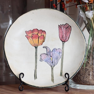 May include: A round, off-white ceramic plate with a hand-painted floral design. The plate features three tulips in shades of orange, pink, and purple. The rim has a green and black trim. The plate is displayed on a black metal stand.