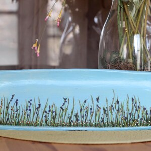 May include: A rectangular, light blue ceramic tray with a hand-painted design of tall green grass, purple flowers, and small gold accents. The tray is displayed on a light brown surface, with a glass vase in the background.