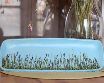 May include: A rectangular, light blue ceramic tray with a hand-painted design of tall green grass, purple flowers, and small gold accents. The tray is displayed on a light brown surface, with a glass vase in the background.