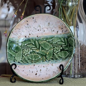 May include: A ceramic plate with a cream-colored rim and a central band of green leaves and berries. The plate is displayed on a black metal stand. The plate's design features a textured, embossed pattern.