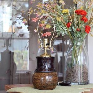 May include: A decorative table lamp with a dark brown ceramic base and a gold-colored metal top. The lamp is next to a clear glass vase filled with colorful flowers. The lamp's cord is visible.