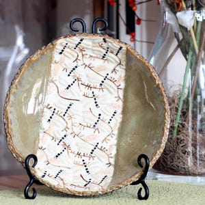 May include: A decorative, round serving bowl with a cream and beige patterned design, displayed on a black metal stand. The bowl has a textured, gold-toned rim and a glossy finish. A clear glass vase with flowers is in the background.