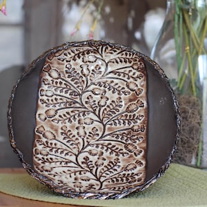 May include: A handmade ceramic plate with a brown rim and a central design featuring a floral pattern in cream and brown tones. The plate has a rustic, textured appearance, suitable for serving food or as a decorative piece.