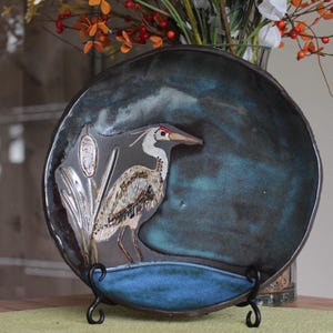 May include: A decorative ceramic plate featuring a heron design. The plate has a dark blue and gray background with a raised image of a heron in shades of white, brown, and gray. The plate is displayed on a black metal stand.