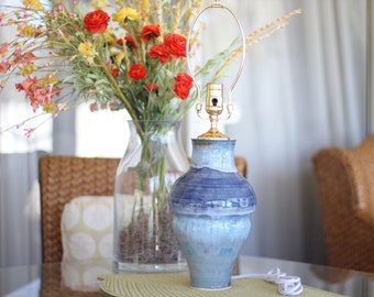 May include: A blue and white ceramic table lamp with a gold metal base and shade holder. The lamp is sitting on a table with a green placemat. There are flowers in a vase behind the lamp.