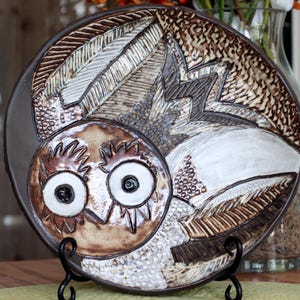 May include: A decorative ceramic plate featuring an owl design. The plate has a brown, white, and tan color scheme with textured details. The owl's face is centered, with large eyes and a detailed feather pattern. The plate is displayed on a black metal stand.