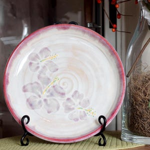 May include: A decorative ceramic plate with a white and pink color scheme. The plate features a floral design in shades of purple and yellow. It is displayed on a black metal stand, suggesting its use as a decorative item or for serving.
