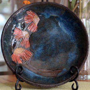 May include: A round ceramic plate featuring a deep blue and black mottled glaze. The surface is decorated with three raised, textured orange flowers with yellow centers arranged in a vertical line along the left side of the plate.