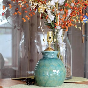 May include: A teal ceramic lamp base with a glossy, mottled glaze and a rounded, bulbous shape. It features a gold-toned metal lamp harp and socket assembly attached to the top. The surface has a smooth, reflective finish with subtle color variations.