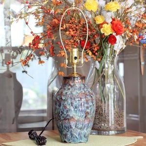 May include: A ceramic table lamp base with a glossy, multi-colored drip glaze in shades of blue, brown, and cream. The lamp features a gold-toned metal socket and a harp attachment, with a black electrical cord extending from the base.