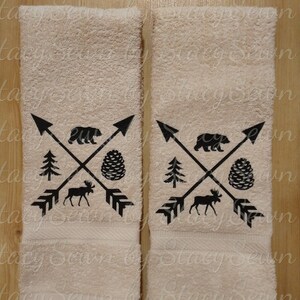 May include: Two beige hand towels with black embroidered designs. The design features a moose, a bear, a pine cone, and arrows.