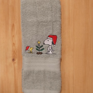 May include: A gray hand towel with an embroidered Christmas design. The design features Snoopy and Woodstock wearing Santa hats next to a small Christmas tree. The towel is made of a soft, absorbent material.