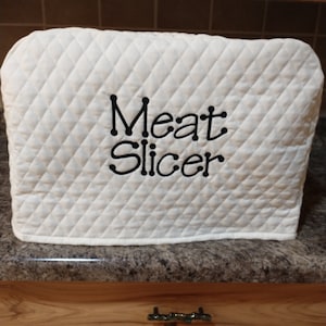 May include: White quilted appliance cover with black embroidery that reads "Meat Slicer".