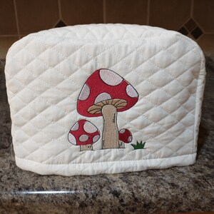 May include: White quilted toaster cover with an embroidered design of red and white mushrooms with brown stems and green grass.