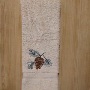 May include: Cream-colored bath towel with an embroidered pine cone and pine needles. The towel is folded and has a decorative border. The embroidery features shades of brown and green.