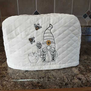 May include: White quilted toaster cover with an embroidered gnome holding a honey dipper and a jar of honey. Two bees are flying around the gnome.