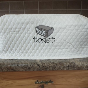 May include: White quilted toaster cover with a diamond pattern. The cover features an embroidered image of a toaster and the word "toast" in black script. The cover is designed to protect a toaster from dust and debris.