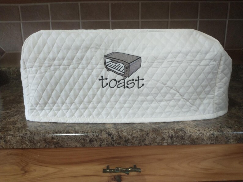 May include: White quilted toaster cover with a diamond pattern. The cover features an embroidered image of a toaster and the word "toast" in black script. The cover is designed to protect a toaster from dust and debris.