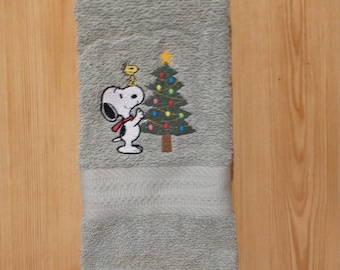 May include: A light green hand towel featuring an embroidered image of Snoopy wearing a red scarf and Woodstock next to a decorated Christmas tree. The towel is folded and has a textured appearance.