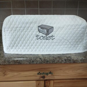 May include: White quilted toaster cover with a diamond pattern. The cover features an embroidered image of a toaster and the word "toast" in a script font. The cover is designed to protect a toaster from dust and debris.