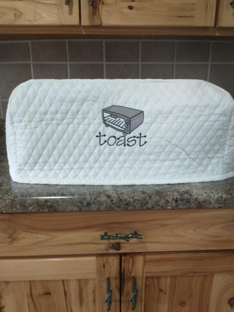 May include: White quilted toaster cover with a diamond pattern. The cover features an embroidered image of a toaster and the word "toast" in a script font. The cover is designed to protect a toaster from dust and debris.