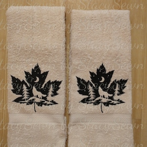 May include: Two beige hand towels with a black embroidered design of a wolf howling at the moon in a forest scene inside a maple leaf.