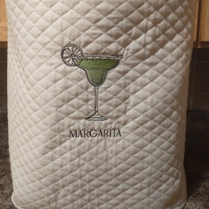 May include: A cream-colored quilted appliance cover featuring an embroidered green margarita cocktail in a stemmed glass with a lime slice garnish. The word MARGARITA is embroidered in black capital letters directly below the cocktail graphic.