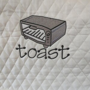 May include: White quilted fabric with a stitched design of a gray toaster oven. The word "toast" is embroidered in black script below the oven. The fabric has a diamond pattern.