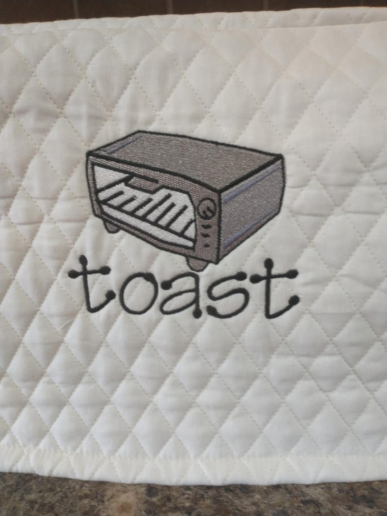 May include: White quilted fabric with a stitched design of a gray toaster oven. The word "toast" is embroidered in black script below the oven. The fabric has a diamond pattern.