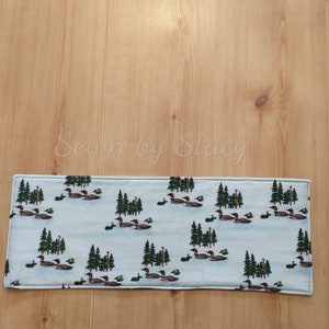 May include: A white fabric table runner with a repeating pattern of brown ducks swimming in a blue lake with green pine trees. The text "Sewn by Stacy" is visible in the background.