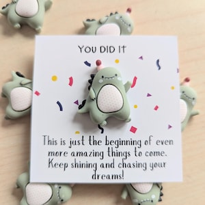 Puede incluir: Una tarjeta de felicitación con el texto "YOU DID IT" y un pin de dinosaurio de dibujos animados. La tarjeta está rodeada de varios pines de dinosaurios verdes con sombreros de fiesta. La tarjeta también tiene el texto: "This is just the beginning of even more amazing things to come. Keep shining and chasing your dreams!"