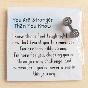 May include: A white card with the text "You Are Stronger Than You Know" and an encouraging message. A small, silver dumbbell pin is attached to the card, adding a motivational touch. The card is in a clear plastic sleeve.