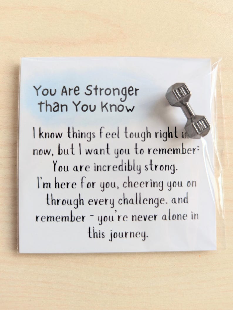 May include: A white card with the text "You Are Stronger Than You Know" and an encouraging message. A small, silver dumbbell pin is attached to the card, adding a motivational touch. The card is in a clear plastic sleeve.