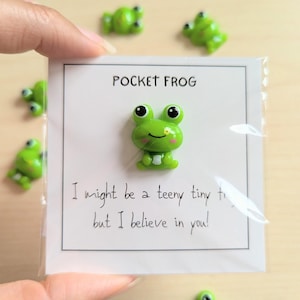 Peut inclure: Une figurine de grenouille verte avec de grands yeux noirs et un ventre blanc est présentée sur une carte blanche. La carte indique "POCKET FROG" et "I might be a teeny tiny but I believe in you !" Plusieurs autres figurines de grenouilles sont éparpillées.