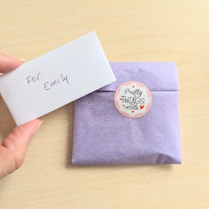 May include: A small, wrapped package in lavender tissue paper, sealed with a round sticker that reads "Pretty Things Inside." A handwritten note that says "For: Emily" is held nearby.