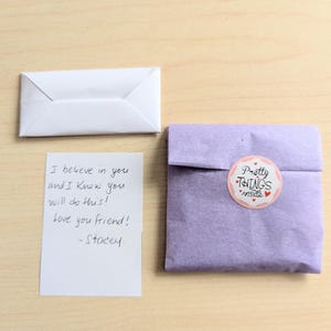 May include: A white folded envelope, a handwritten note, and a lavender package with a sticker that reads "Pretty Things Inside" are arranged on a light wood surface. The note says "I believe in you and I Know you will do this! Love you friend! -Stacey."