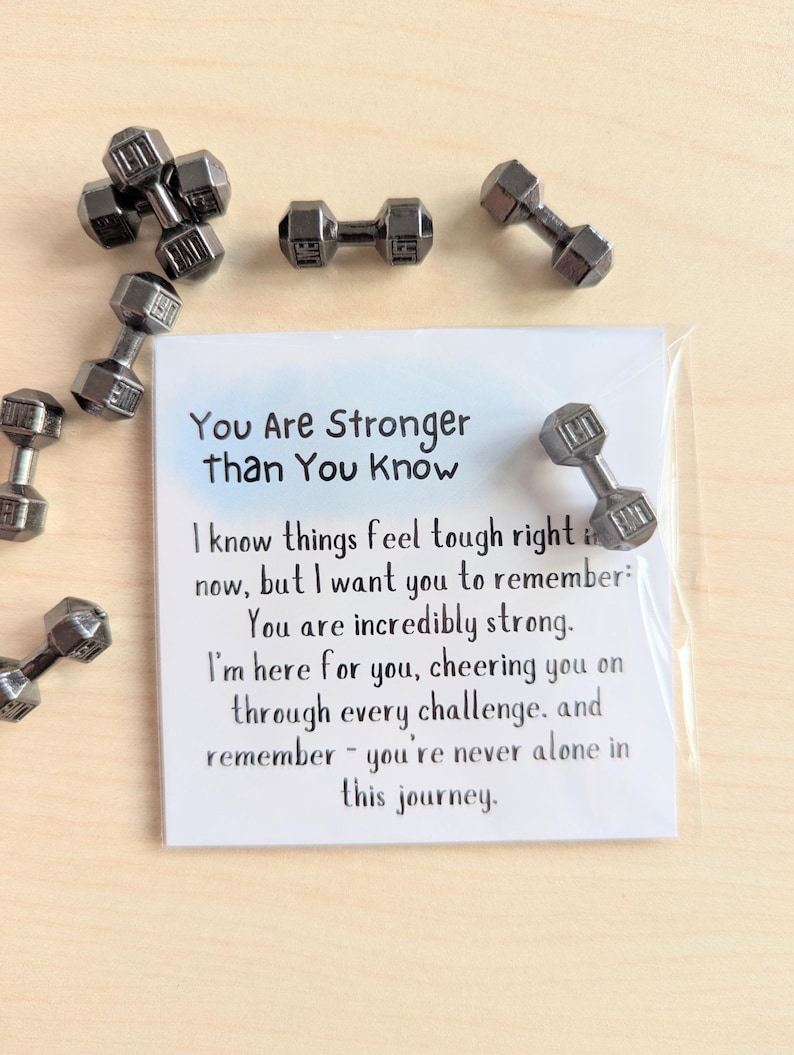 May include: Small, silver-colored dumbbell-shaped magnets scattered around a white card. The card has the text "You Are Stronger than You Know" and an inspirational message. The dumbbells appear to be made of metal.