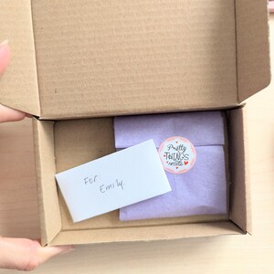 May include: An open cardboard box reveals a wrapped gift and a handwritten note. The gift is wrapped in lavender paper and sealed with a sticker that reads "Pretty Things inside." The note says "For: Emily."