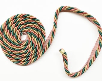 May include: A close-up of a green, gold, and pink braided trim. The trim is coiled into a circle and a long piece is extending from the circle.