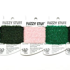 May include: Five spools of Fuzzy Stuff yarn in various colors: two shades of green, a dark pink, and a dark green. Each spool is labeled with the brand and product information. The yarn is fluffy and textured, ideal for craft projects.