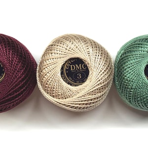 May include: Three balls of DMC size 3 cotton embroidery thread in deep burgundy, light beige, and sage green. Each ball features a black circular label with the text DMC, 3, and Made in France, indicating 50g and 147m of thread.
