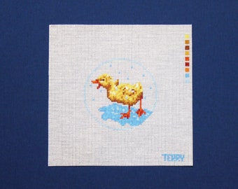 May include: A yellow duckling with a blue background, surrounded by white dots, is depicted in a cross-stitch pattern on a white canvas.