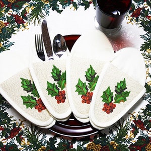 May include: Four white fabric place card holders with a holly and berry design. The holders are arranged in a circle on a red and green tablecloth.