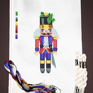 May include: A white canvas with a colorful nutcracker design. The nutcracker is wearing a red and blue uniform with a gold belt and a black hat. The nutcracker is holding a sword in its right hand. There are three spools of white embroidery floss on the right side of the canvas.