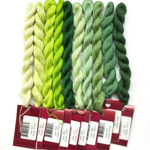 May include: A collection of green embroidery floss skeins in various shades, from light to dark. Each skein is labeled with a red tag, displaying color and dye lot information. The floss is arranged vertically against a white background.