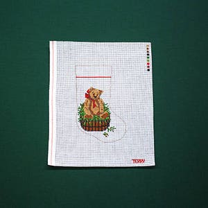May include: White canvas stocking with a teddy bear sitting in a basket with green leaves and a red bow. The stocking has a red line across the top and the word "Teddy" printed at the bottom.