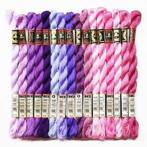 May include: A collection of 15 spools of DMC embroidery floss in shades of purple, lavender, and pink. The spools are arranged in a row, with the numbers 211, 554, 553, 552, 550, 341, 340, 333, 327, 316, 605, 604, 603, 3689, 3688, and 3687 visible on the labels.