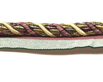 May include: Close-up of decorative trim featuring a twisted rope design in shades of beige, green, and purple. Below the rope is a woven fabric with a white background, a pink stripe, and a green scalloped edge.