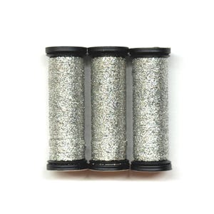 May include: Three spools of silver glitter thread with black caps and bases are arranged vertically against a white background. The thread is tightly wound, creating a textured, sparkling appearance. The spools are evenly spaced.
