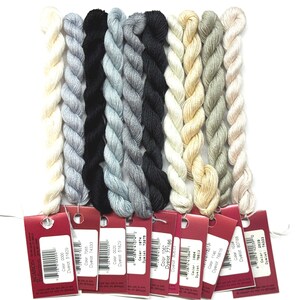 May include: A collection of embroidery floss in various colors, including cream, gray, black, and beige. Each skein is labeled with a color code and brand information. The floss is neatly arranged, showcasing a range of neutral and muted tones.